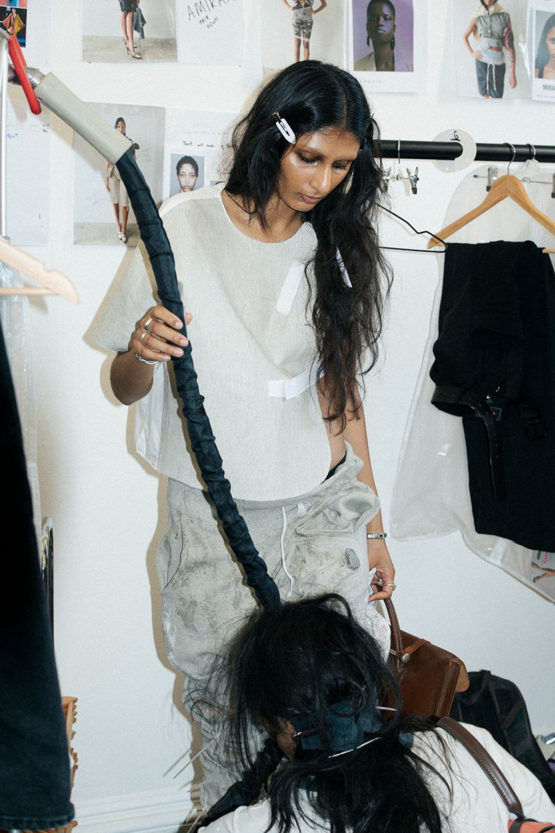 Woman in loose-fitting top standing in dressing room with another person in a chair; clothing rack and photos in the background.