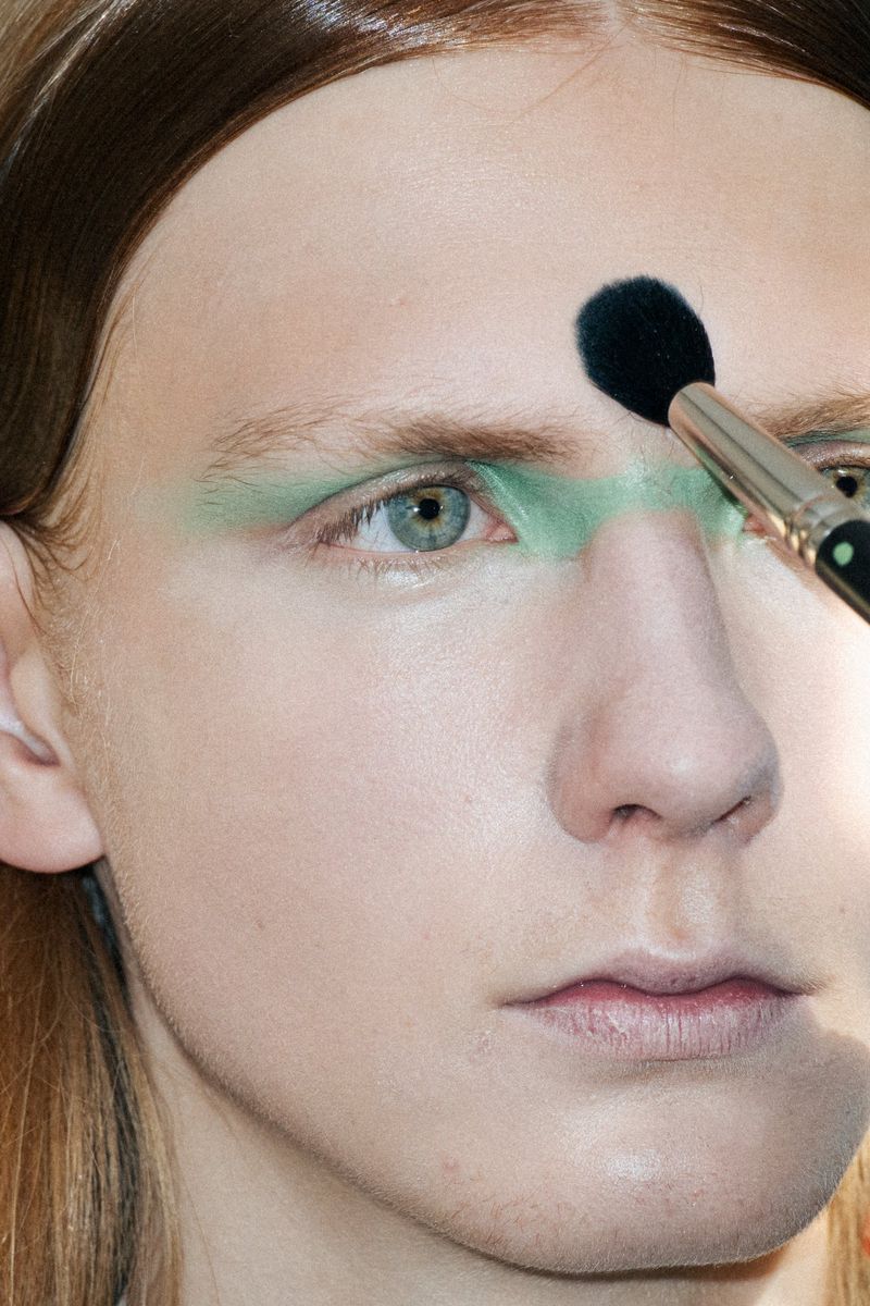 Close-up of a person getting green makeup applied to their face with a makeup brush in a beauty or fashion setting.
