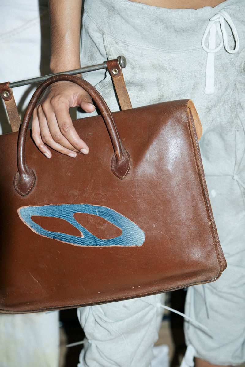 Person holds a brown leather satchel bag with a blue design, accessorized with a tan strap and silver metal hardware.