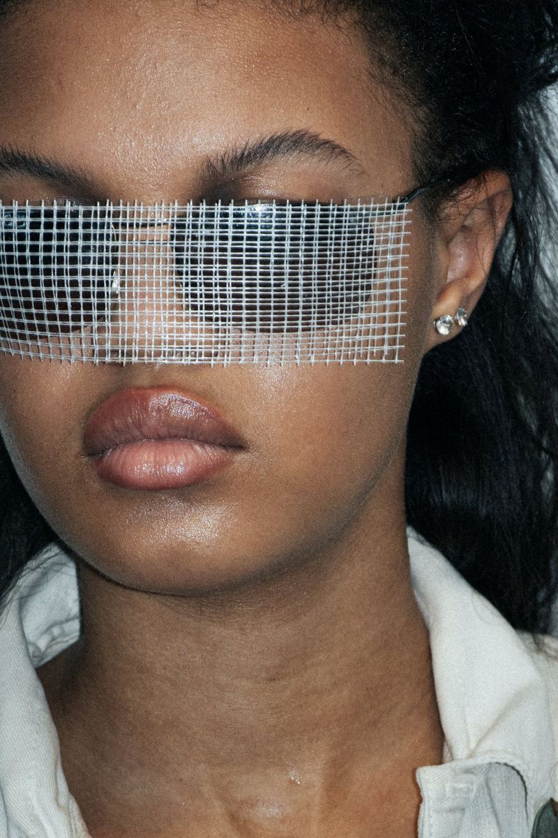 Close-up shot of a stylish adult woman with dark hair, wearing a light-colored jacket and grid-pattern eyewear against a dark backdrop.