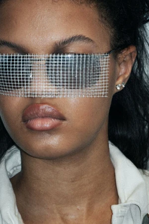 Close-up shot of a stylish adult woman with dark hair, wearing a light-colored jacket and grid-pattern eyewear against a dark backdrop.