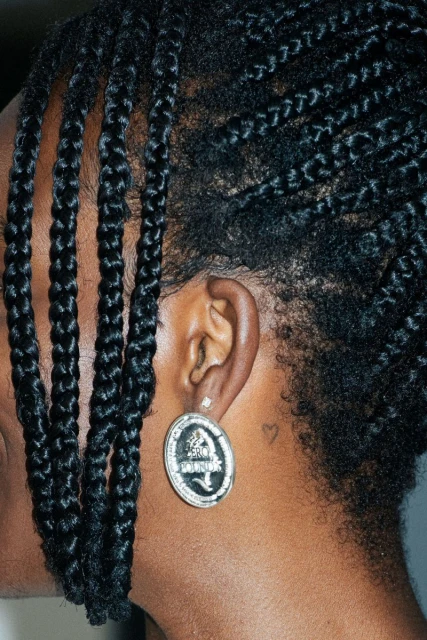 Close-up studio shot of a black woman's ear with an ornate silver earring, showcasing braided hair, and a minimalist heart tattoo on her neck.
