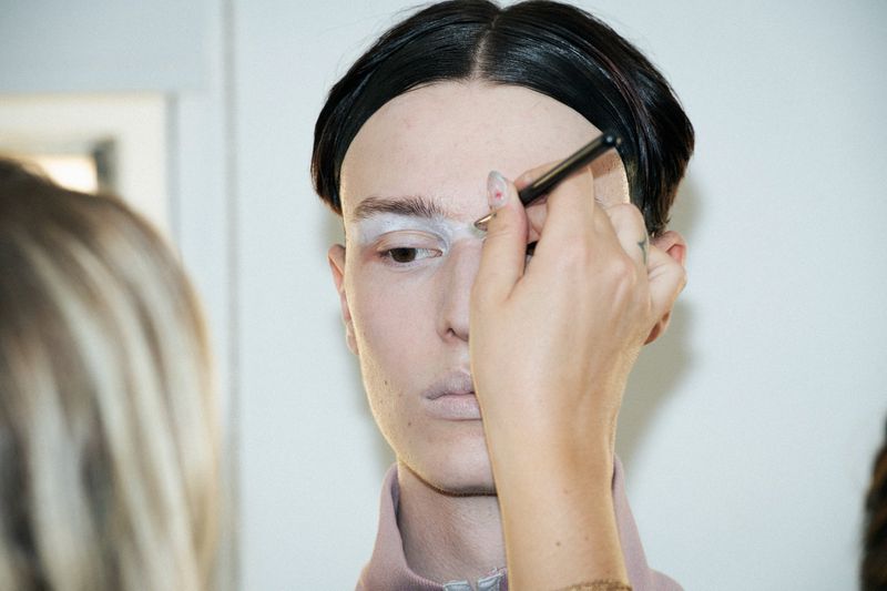 An adult person with makeup being applied with a makeup brush in an indoor setting, captured in a beauty or fashion editorial style.