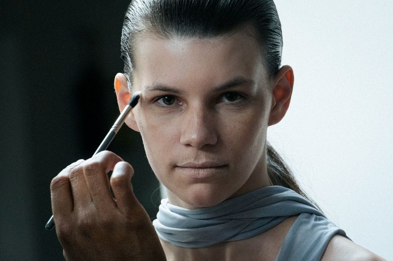 Close-up shot of an adult female model having makeup applied with a brush. Focus on the model's face in a muted colour palette.