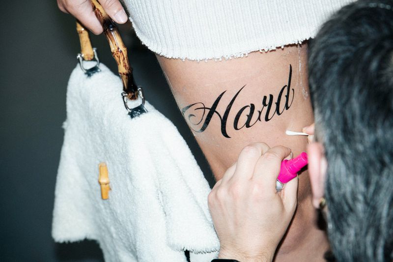 Close-up of a leg with temporary tattoo, artist applying makeup with a cotton swab, bamboo-handled towel nearby against a grey background.
