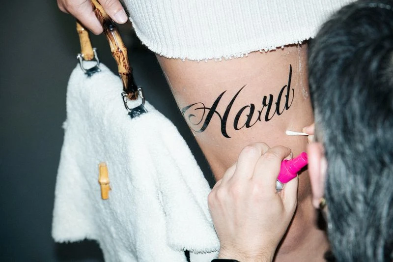Close-up of a leg with temporary tattoo, artist applying makeup with a cotton swab, bamboo-handled towel nearby against a grey background.