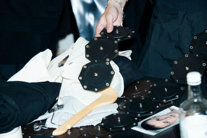 Close-up of clothing items, a belt composed of black disks, a wooden hanger, and a portrait photo on a dark surface. 