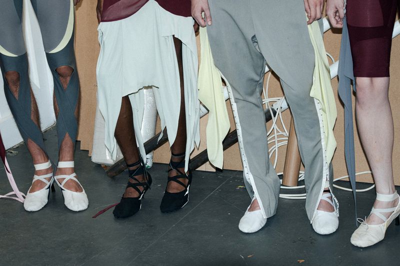 Fashion runway show with models showcasing avant-garde clothing and unique footwear in a neutral setting, legs and feet only, bright lighting.