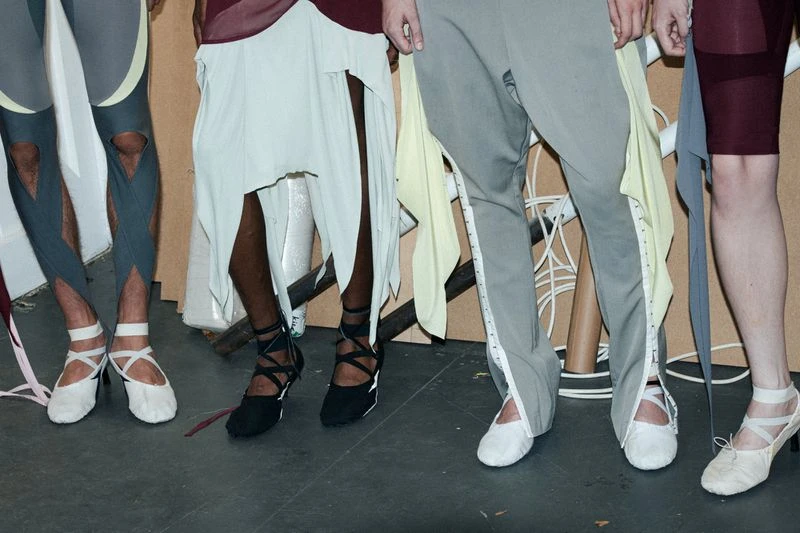 Fashion runway show with models showcasing avant-garde clothing and unique footwear in a neutral setting, legs and feet only, bright lighting.