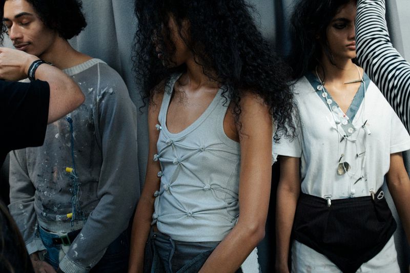 Three models showcase unique fashion designs indoors, with an individual adjusting the shirt of the model on the left side of the frame.