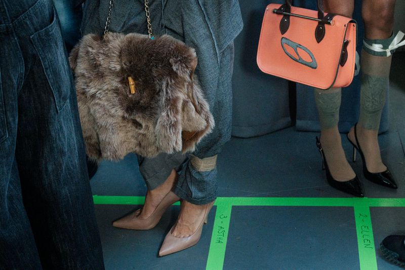 Models backstage at a fashion show, showcasing stylish fur and leather purses, along with elegant high heels and fashionable patterned stockings.