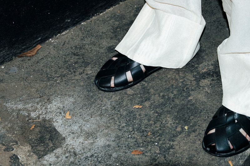 Close-up of a person wearing woven leather sandals and light-colored linen pants on a concrete surface with black wall in background.