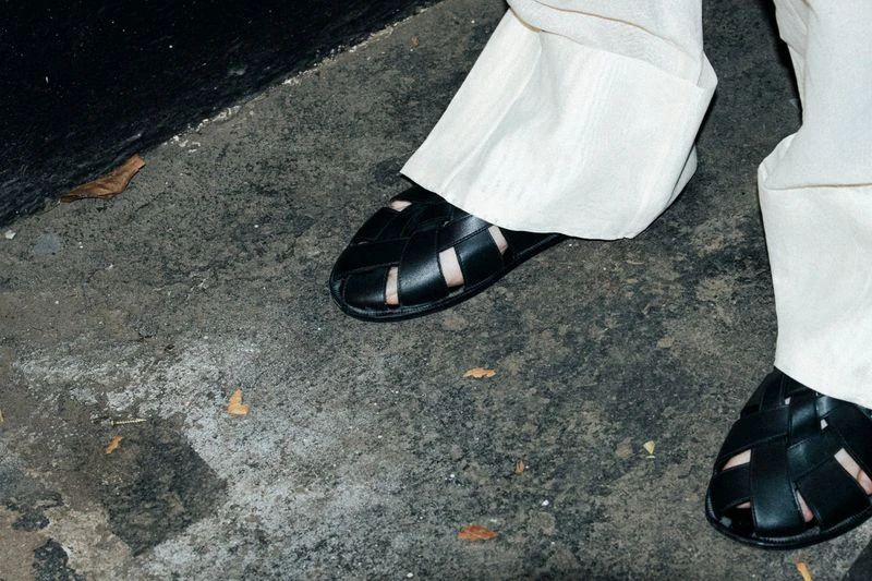 Close-up of a person wearing woven leather sandals and light-colored linen pants on a concrete surface with black wall in background.