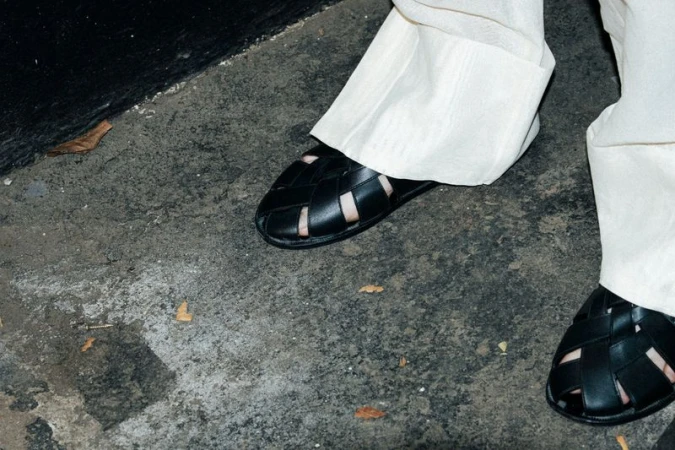 Close-up of a person wearing woven leather sandals and light-colored linen pants on a concrete surface with black wall in background.