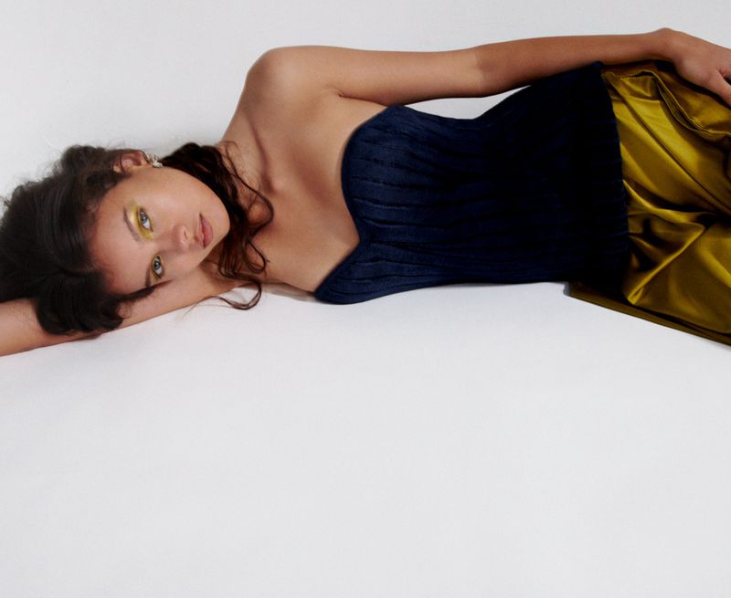 Woman lying down in a blue corset top and gold skirt against a white background. She has gold eye makeup and dark wavy hair.