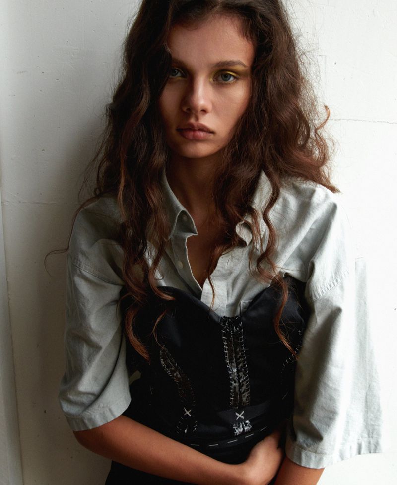 Portrait of a woman with wavy brown hair wearing a light short-sleeve shirt layered under a black corset against a white backdrop.
