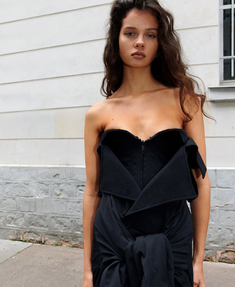 A woman with long brown hair models a strapless black dress with architectural detailing against a white-painted brick wall backdrop.