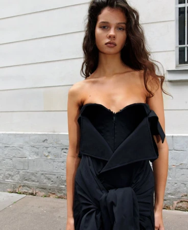 A woman with long brown hair models a strapless black dress with architectural detailing against a white-painted brick wall backdrop.