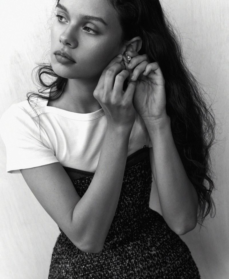 Portrait of a young woman with long brown hair adjusting an earring and looking off to the side; she wears a white T-shirt and dark dress.