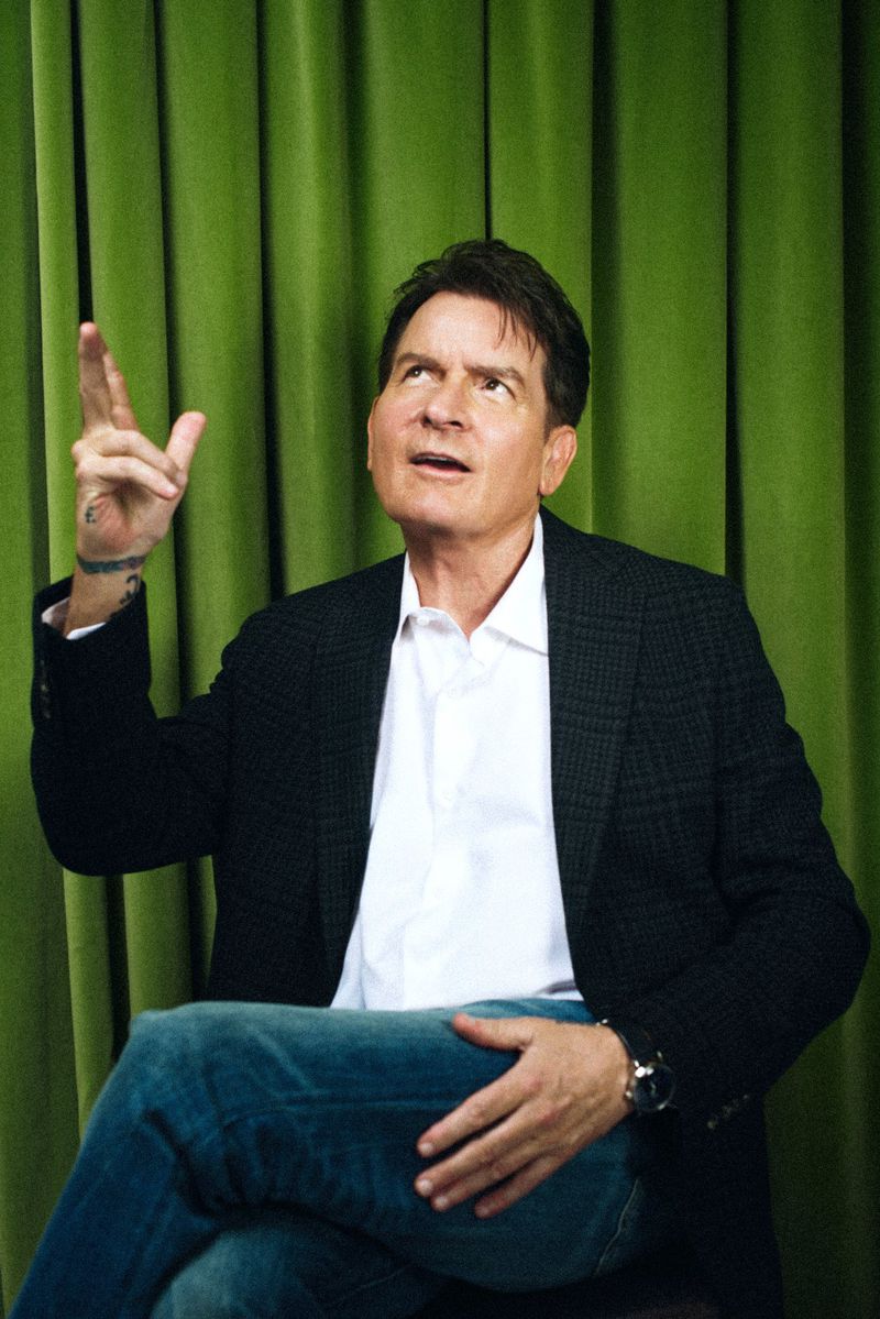 Portrait of actor Charlie Sheen, wearing a white shirt and blazer, posing in front of a green curtain while pointing up.
