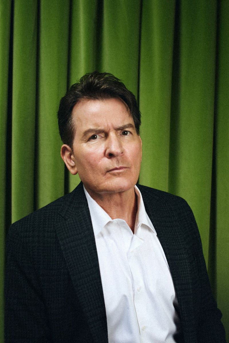 Portrait of adult male, Charlie Sheen, wearing a white collared shirt and black blazer, gazing into the camera against green curtain backdrop.