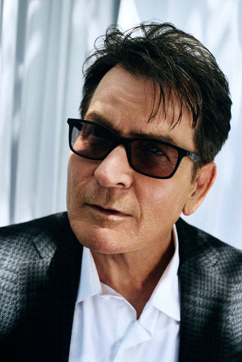 Headshot of a mature adult male with dark hair wearing a white shirt, blazer, and dark glasses against a blurry, light background.