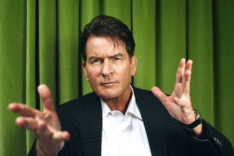 Portrait of Charlie Sheen in a suit jacket with hands up, standing against a green curtain backdrop. The image is well-lit and focused.