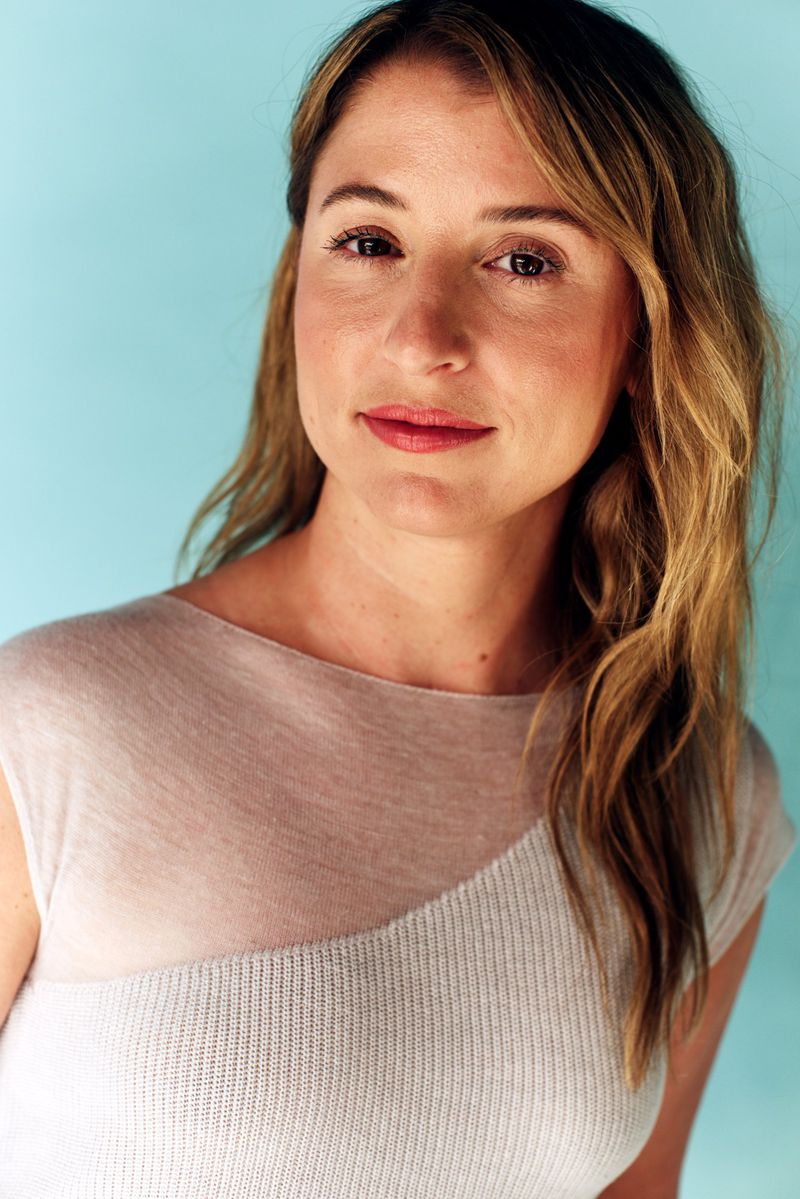 Headshot of a smiling woman with long brown hair wearing a light-colored top against a light blue background in an indoor setting.