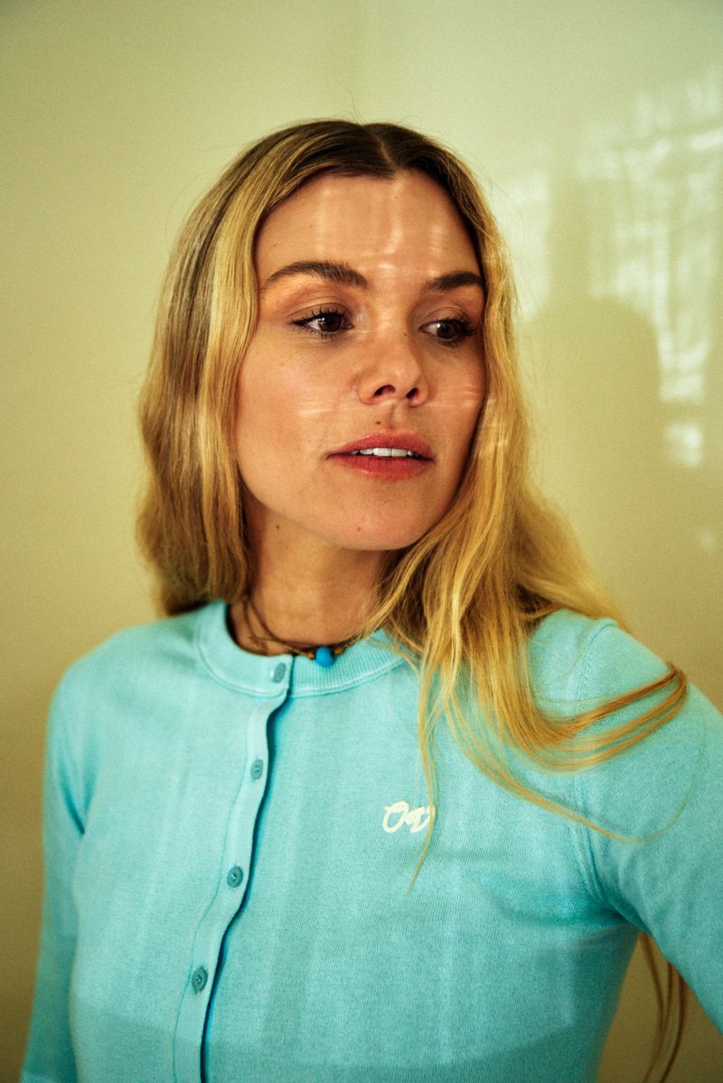 Blonde woman in a light blue button-down shirt looks off to the side in a studio with soft, diffused lighting and light shadow patterns.