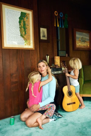 A woman sits on the floor with a child, with another child holding a guitar nearby, in a wood-paneled room with a map on the wall.