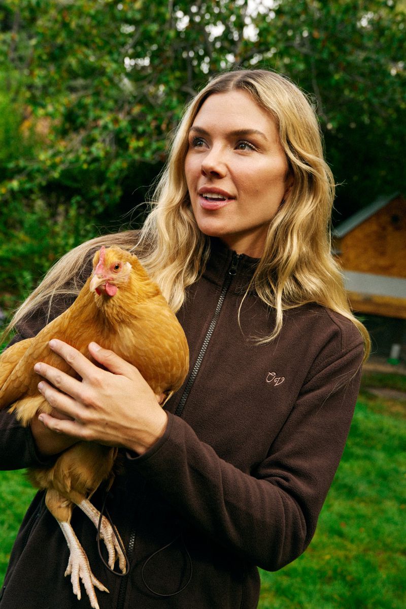 Blonde woman in a brown jacket holding a brown chicken outdoors with a green background and a partial view of a brown building.