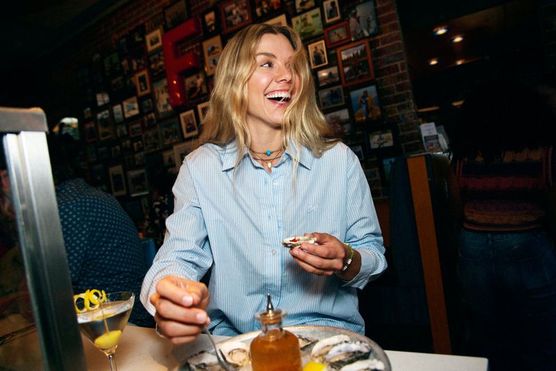 A blonde woman in a light blue striped shirt laughs while holding an oyster in a dimly lit restaurant. A martini and seafood platter are visible.