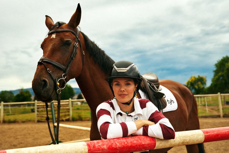 Woman wearing a riding helmet and striped shirt poses with a brown horse in an outdoor paddock with a red and white rail fence.