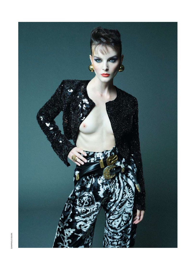 A fashion shot of an adult female with a bold hairstyle wearing a sparkly black jacket, patterned pants, jewelry, and posing against a dark gray backdrop.