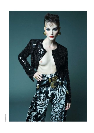 A fashion shot of an adult female with a bold hairstyle wearing a sparkly black jacket, patterned pants, jewelry, and posing against a dark gray backdrop.