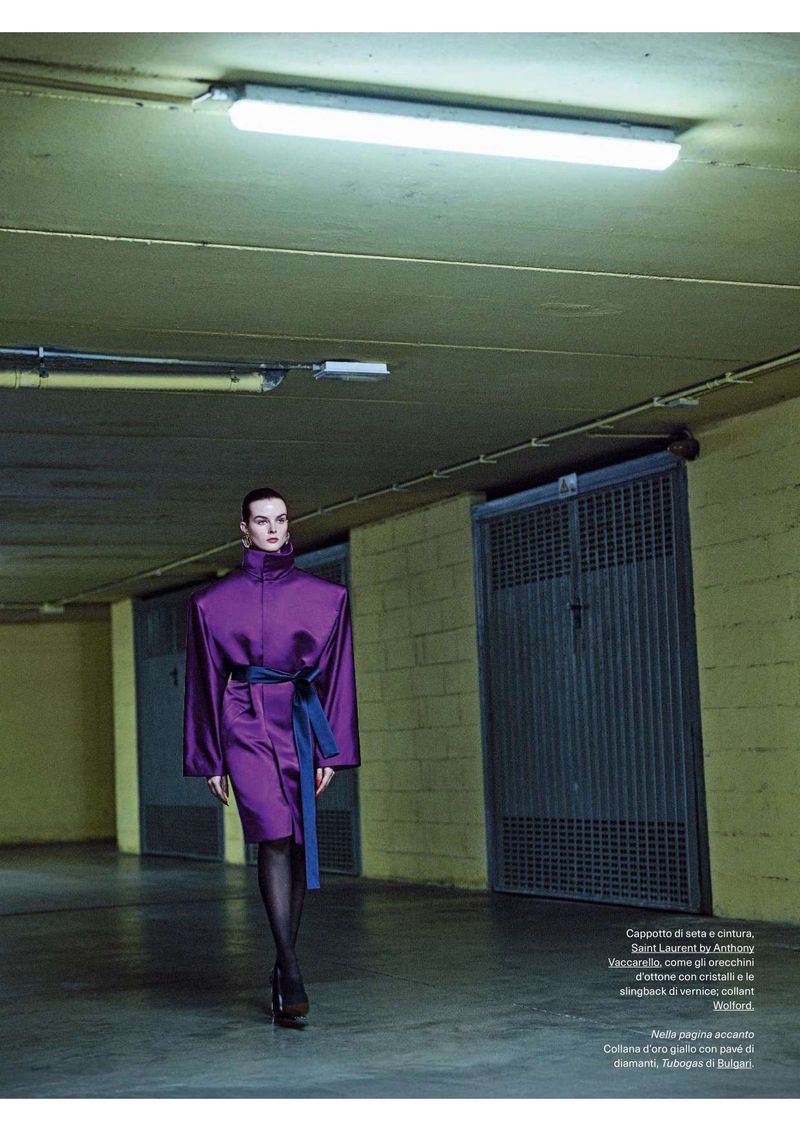 Fashion editorial image of a woman walking in a purple dress with a blue belt, posing in an indoor underground parking garage.