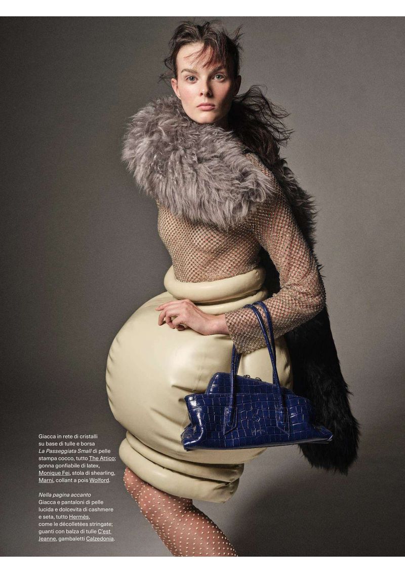 A woman models a fur-trimmed coat and a leather bag in a fashion editorial shot against a grey backdrop. The lighting is soft.