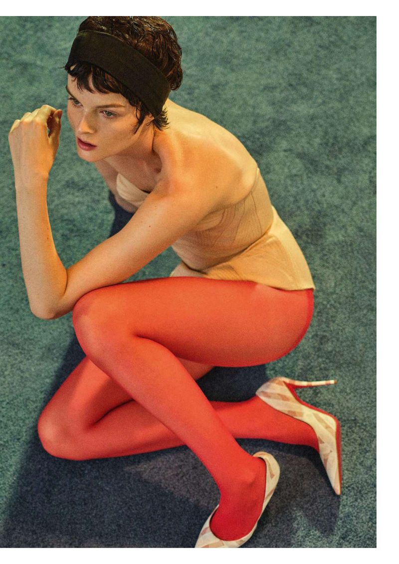 Brunette woman with a black headband, wearing a peach-colored top and red tights, posing in a studio with a blue textured carpet.