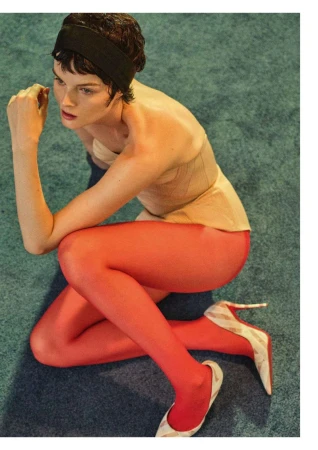Brunette woman with a black headband, wearing a peach-colored top and red tights, posing in a studio with a blue textured carpet.