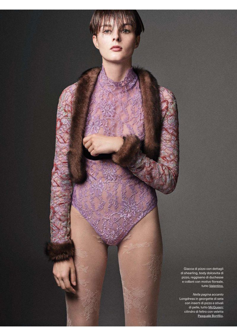 Fashion editorial shot of a model wearing a lilac lace bodysuit, patterned sheer tights, and a dark fur stole in a studio setting.