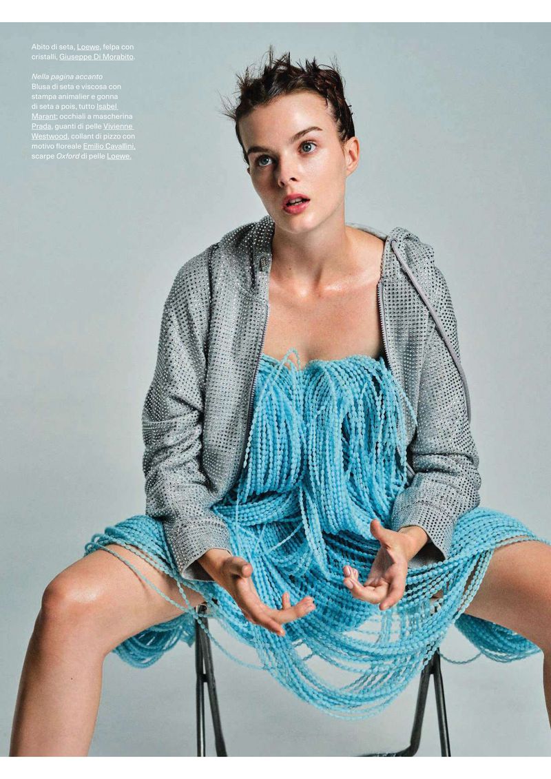 Fashion shot of a young fair-skinned woman with short dark hair wearing an aquamarine beaded dress and grey hoodie, seated indoors.