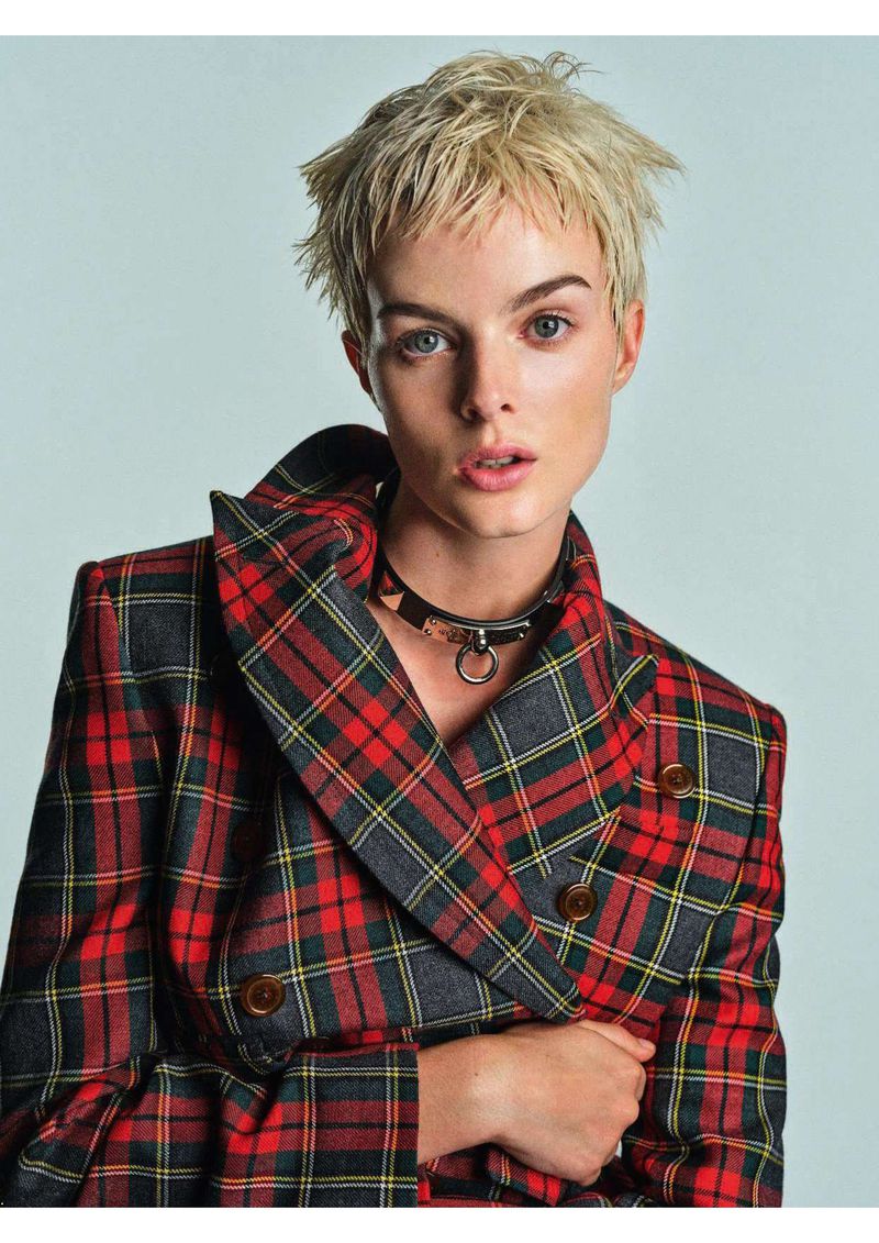 Portrait of a woman with short blonde hair wearing a plaid jacket and black choker against a light gray studio backdrop.