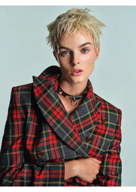 Portrait of a woman with short blonde hair wearing a plaid jacket and black choker against a light gray studio backdrop.