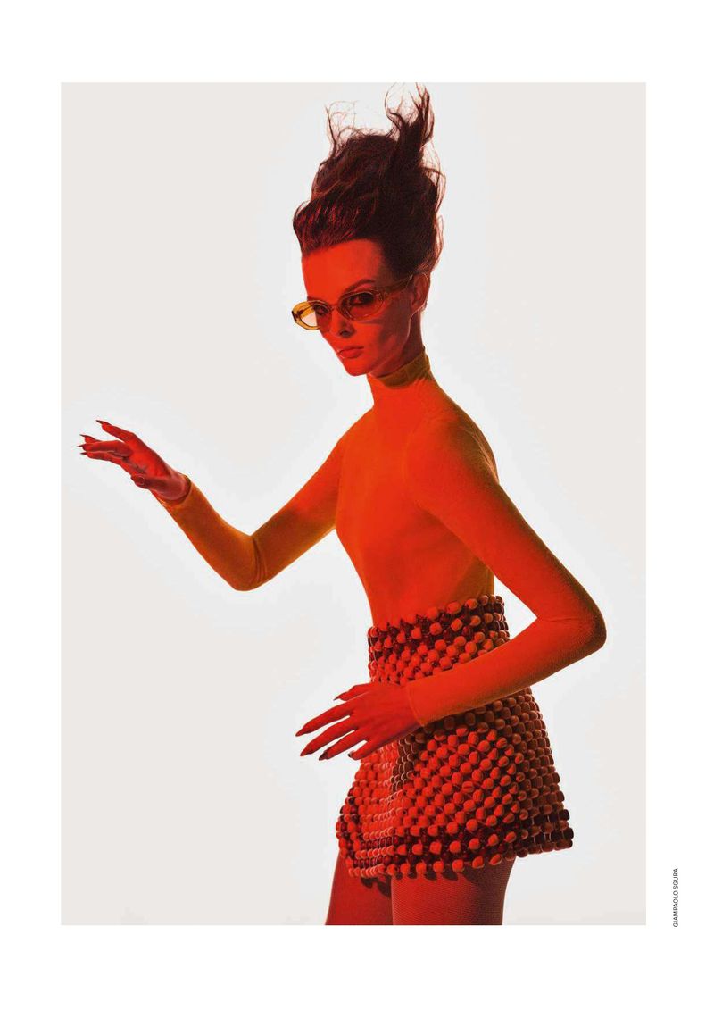 Woman with high hair in orange turtleneck, beaded mini skirt, and sunglasses poses in a fashion photography studio against a white background.