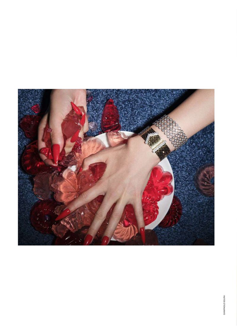 Overhead studio shot showcases two fair-skinned hands with red nails arranging red and pink jelly candies on a white plate over blue carpet.