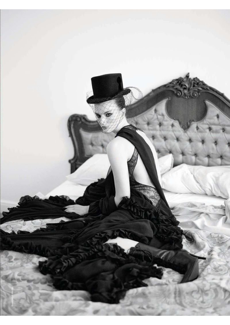 Woman in black hat and gown sits on a bed with pillows. The shot is a black and white studio image with a vintage feel.