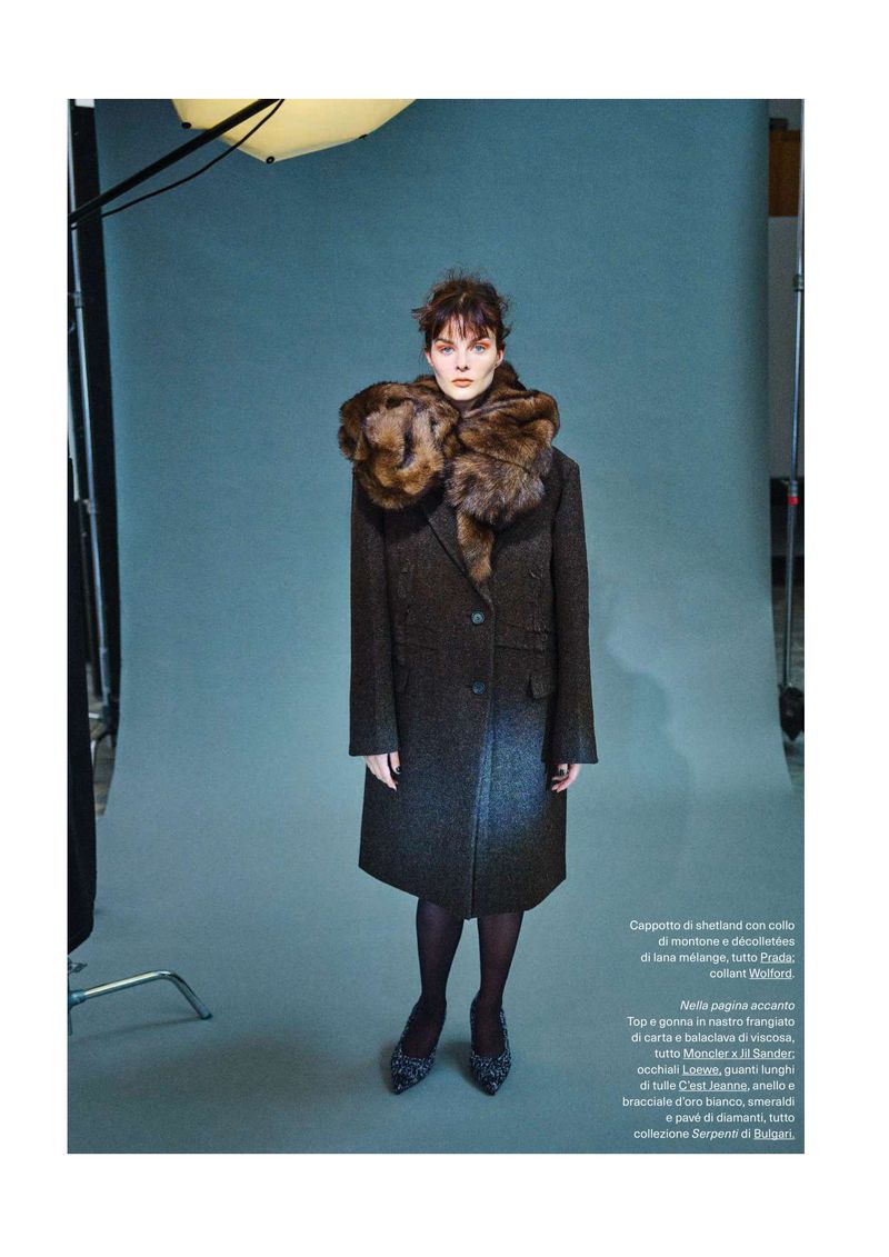 A fashionable woman with brown hair wearing a long brown coat with fur collar and patterned heels in a studio environment with grey backdrop.