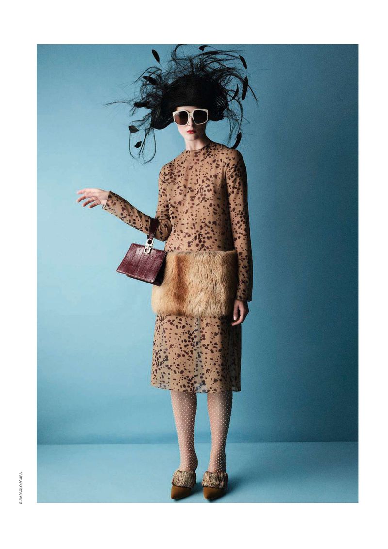 A woman in a polka dot dress and feathered hat poses in front of a blue background, holding a purse in a studio shot.