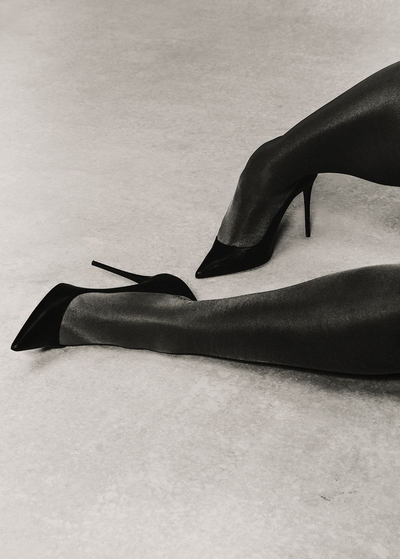 A grayscale fashion shot of an adult woman's legs and black stiletto high heels, set against a simple, light-grey studio backdrop.
