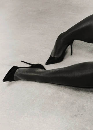 A grayscale fashion shot of an adult woman's legs and black stiletto high heels, set against a simple, light-grey studio backdrop.